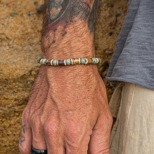 May include: A beaded bracelet with brown, blue, and tan beads. The bracelet is worn on a wrist with a tattoo. A black ring is on the finger. The person is wearing a gray shirt and tan pants.