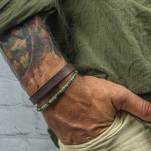 Op de afbeelding: Close-up van twee armbanden om de pols van een persoon. De bovenste armband is een donkerbruine leren band. De tweede armband is gemaakt van kleine, ronde, groene kralen. De persoon draagt een groen overhemd en een kaki broek.