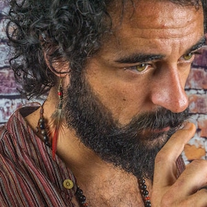 Könnte beinhalten: Ein Mann mit langen, lockigen dunklen Haaren und einem dichten Bart trägt ein gestreiftes Hemd mit einer Perlenkette. Er trägt einen schwarzen Ring am Finger und einen Federschmuck am linken Ohr.