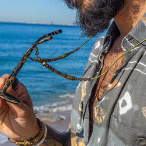 Op de afbeelding: Zonnebril met een patroonmontuur en een kralenketting. De zonnebril wordt vastgehouden voor een overhemd met patroon en een button-down kraag. De achtergrond toont de oceaan en een zandstrand.