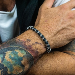 Op de afbeelding: Zwart kralenarmband met zilverkleurige accenten. De armband heeft donkere, ronde kralen afgewisseld met zilverkleurige cilindrische kralen. De sieraden worden gedragen om een pols met zichtbare tatoeages.