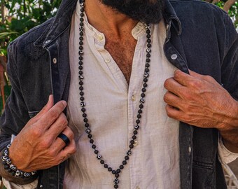 May include: A black beaded necklace and bracelet with white detailing. The long necklace has a pendant. The person is wearing a white shirt and a dark denim jacket. The background is a natural outdoor setting with plants.