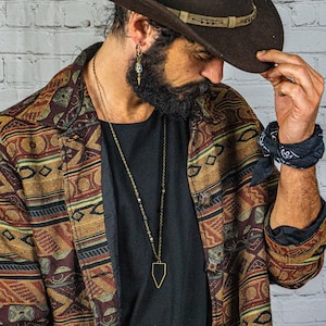 Könnte beinhalten: Eine Person trägt einen braunen Cowboyhut, ein schwarzes T-Shirt, eine gemusterte braune Jacke und eine schwarze Halskette mit einem Schild-Anhänger. Die Person trägt ein schwarzes Bandana am Handgelenk.