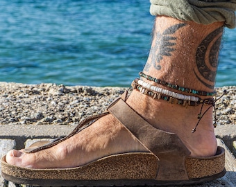 May include: A person's ankle with a tattoo of a bird and a crescent moon, wearing a brown leather sandal and multiple beaded anklets.