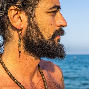 Puede incluir: Un hombre con el pelo largo, oscuro y rizado y barba lleva un collar largo de cuentas y un solo pendiente con múltiples cuentas y abalorios. Está de pie frente a una masa de agua azul.