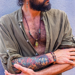 May include: A man wearing a green shirt with a brown leather cord necklace with a gold pendant and colorful beads. He is wearing a brown and gold beaded bracelet on his wrist.
