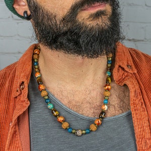 Op de afbeelding: Een veelkleurige kralenketting met blauwe, oranje en bruine kralen. De ketting wordt om de nek gedragen. De persoon draagt een oranje corduroy overhemd. Een sieraad.