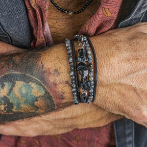 Puede incluir: Una pulsera de capas con cordón de cuero negro, cuentas negras y un dije de plata en la muñeca de un hombre. La pulsera está en un brazo tatuado.