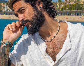 May include: A man with a beard and a straw hat is wearing a white t-shirt and a beaded necklace. He is standing in front of a blue body of water.