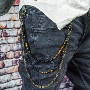 May include: A multi-strand wallet chain with black and gold beads and a gold-colored chain. The chain is attached to a pair of dark gray jeans. The chain is a stylish accessory.