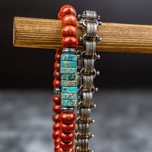 Può includere: Tre braccialetti esposti su un supporto di legno. Un braccialetto presenta perline rosse, turchesi e marroni. Il secondo braccialetto è in metallo argentato con un design intricato. I braccialetti sono esposti su uno sfondo scuro.