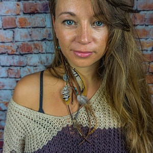 May include: A woman with long brown hair wearing a beige and purple striped jumper with a feather and bead necklace. The background is a brick wall.