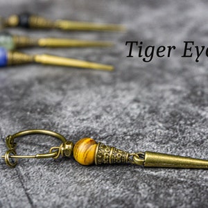 Può includere: Primo piano di orecchini fatti a mano con una perla occhio di tigre e un design in metallo color ottone. Gli orecchini hanno una forma lunga e appuntita. Il testo "Tiger Eye" è visibile nell'immagine. Altri orecchini sono leggermente sfocati sullo sfondo.