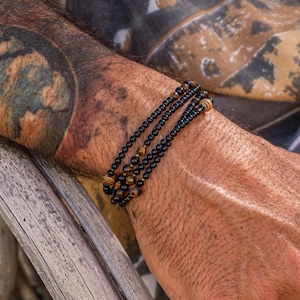 Op de afbeelding: Een armband met meerdere strengen met zwarte kralen en tijgeroogaccenten. De armband wordt gedragen om een pols met een gedetailleerde tatoeage. De persoon draagt een grijs overhemd met patroon.