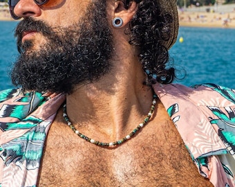 May include: A person wearing a straw hat, sunglasses, and a pink floral shirt, standing near the ocean. The background shows a beach and buildings under a clear blue sky. A beaded necklace is visible.