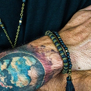 Puede incluir: Pulsera de cuentas negras con detalles dorados y una borla negra. La pulsera se lleva en un brazo con un tatuaje colorido de un globo terráqueo. También se ve un collar de cadena dorada.