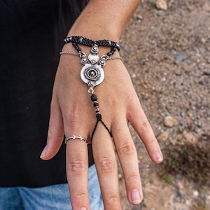 Puede incluir: Una pulsera de cadena de mano plateada con un colgante circular central y detalles de cuentas negras. La pulsera se conecta a un anillo y se muestra en una mano, con un tatuaje visible en el brazo.