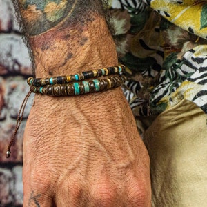 Puede incluir: Dos pulseras de cuentas con detalles marrones, turquesas y dorados se llevan en la muñeca. Las pulseras están apiladas y tienen una estética natural y terrosa. La persona lleva un anillo negro y una camisa floral.