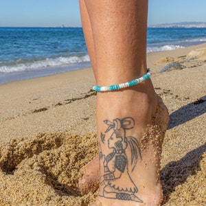 Può includere: Una caviglia di donna con un braccialetto alla caviglia di perline turchesi e bianche. La caviglia ha un tatuaggio di una donna con un vestito. La donna è in piedi su una spiaggia di sabbia con l'oceano sullo sfondo.