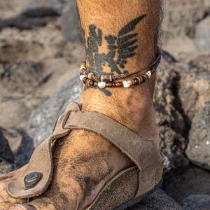 May include: Close-up of a foot wearing a brown leather sandal with a buckle. A beaded ankle bracelet with brown and white beads is worn above the sandal. A tattoo of a winged creature is visible on the leg.