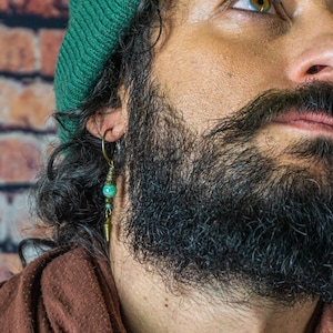Op de afbeelding: Een man met een baard en een groene beanie draagt ​​een turquoise en gouden oorring. De oorring heeft een lang, hangend ontwerp met meerdere kralen.