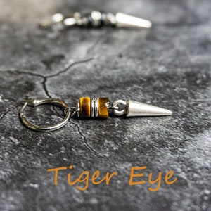 May include: A pair of silver hoop earrings with a tiger eye stone and a silver spike. The earrings are on a gray textured surface. "Tiger Eye" is written in orange text below the earrings.