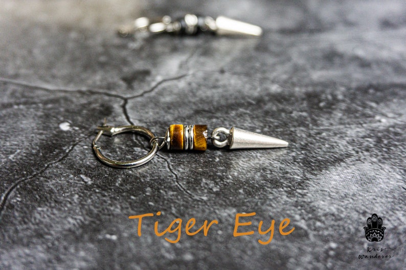 May include: A pair of silver hoop earrings with a tiger eye stone and a silver spike. The earrings are on a gray textured surface. "Tiger Eye" is written in orange text below the earrings.