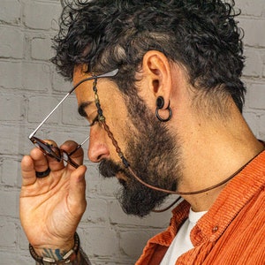 May include: A person wearing a brown corduroy shirt with a beaded glasses chain. The chain is made of brown leather and has beads of various colours and materials. The person is wearing a pair of glasses with brown frames.