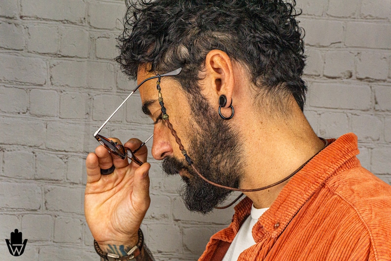 May include: A person wearing a brown corduroy shirt with a beaded glasses chain. The chain is made of brown leather and has beads of various colours and materials. The person is wearing a pair of glasses with brown frames.