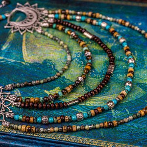 May include: Multi-strand necklace with brown wooden beads, turquoise and green stones, and silver accents. The necklace features two large silver pendants with intricate designs.