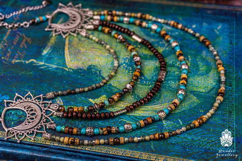 May include: Multi-strand necklace with brown wooden beads, turquoise and green stones, and silver accents. The necklace features two large silver pendants with intricate designs.