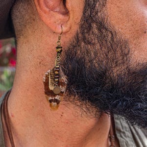 May include: A close-up of a feather earring. The earring has a bronze-colored hook and decorative beads. The feather is brown, white, and tan. A small, round, bronze-colored charm hangs from the earring.
