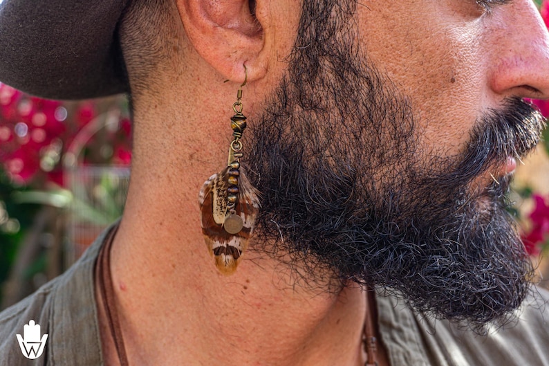 May include: A close-up of a feather earring. The earring has a bronze-colored hook and decorative beads. The feather is brown, white, and tan. A small, round, bronze-colored charm hangs from the earring.