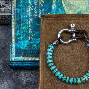 Op de afbeelding: Een turquoise edelsteen armband met een zilveren sluiting en bruine lederen accenten. De armband ligt op een bruine lederen achtergrond met een decoratief patroon.
