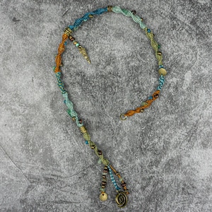 Op de afbeelding: Een veelkleurige kralenketting met een spiraalvormige hanger aan het einde. De ketting is gemaakt van blauw, groen en oranje garen en heeft kleine metalen kralen.