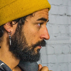 May include: A man with a beard and a yellow beanie is wearing a silver hoop earring with a black bead and a silver spike charm.