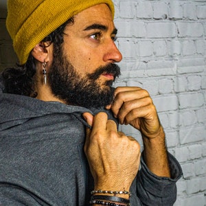 May include: A man wearing a gray hooded sweatshirt with a yellow beanie and silver earrings. He is adjusting the hood of the sweatshirt with his right hand. He is wearing several bracelets on his left wrist.