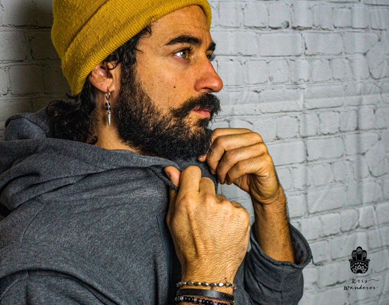May include: A man wearing a gray hooded sweatshirt with a yellow beanie and silver earrings. He is adjusting the hood of the sweatshirt with his right hand. He is wearing several bracelets on his left wrist.