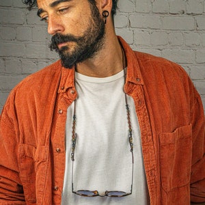 May include: A man wearing a red corduroy shirt with a white t-shirt underneath. He is wearing a beaded glasses chain with glasses hanging from it.