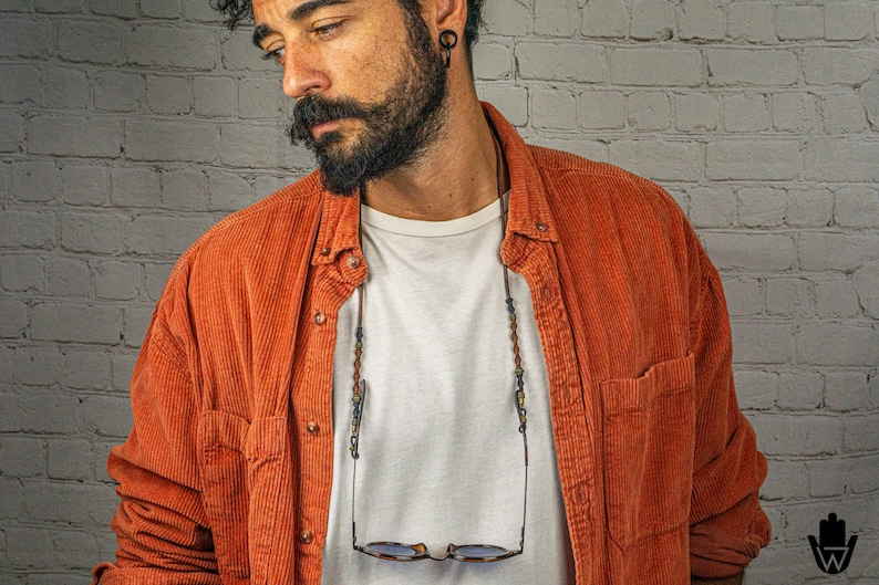 May include: A man wearing a red corduroy shirt with a white t-shirt underneath. He is wearing a beaded glasses chain with glasses hanging from it.