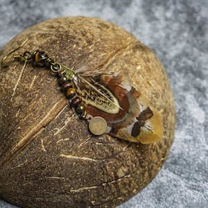 May include: A handcrafted feather earring with a hook closure, featuring a natural feather with brown, tan, and cream hues. The earring is adorned with small beads and a metal charm. The earring rests on a textured brown surface.