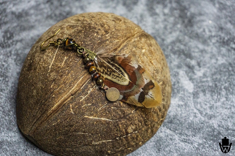 May include: A handcrafted feather earring with a hook closure, featuring a natural feather with brown, tan, and cream hues. The earring is adorned with small beads and a metal charm. The earring rests on a textured brown surface.