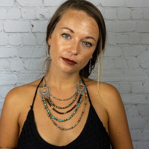 May include: A layered necklace with turquoise, brown, and gold beads, and silver pendants with a sun design. The necklace is worn by a person with long brown hair against a white brick wall.