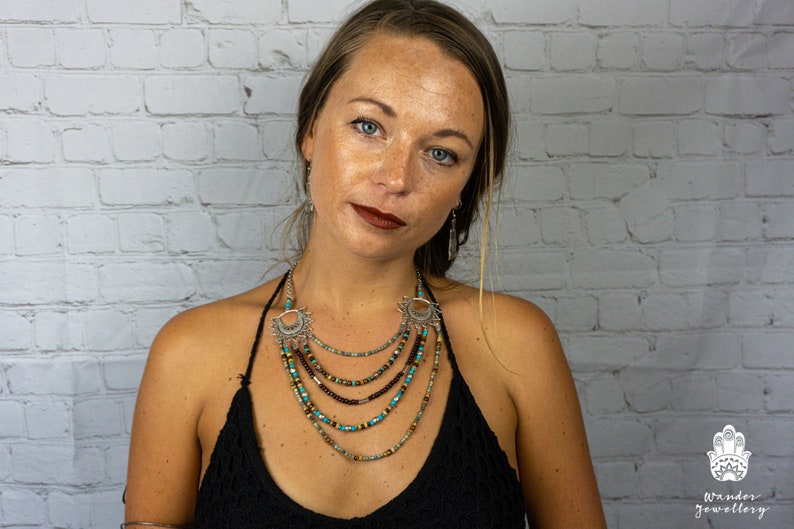 May include: A layered necklace with turquoise, brown, and gold beads, and silver pendants with a sun design. The necklace is worn by a person with long brown hair against a white brick wall.