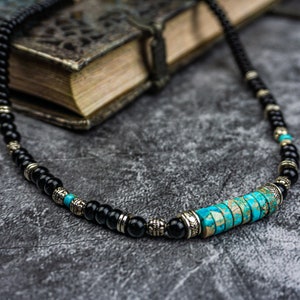 May include: A black beaded necklace with a turquoise and silver centerpiece. The necklace is lying on a gray textured surface.