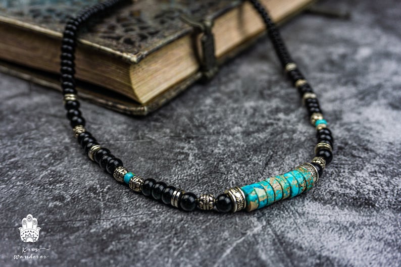 May include: A black beaded necklace with a turquoise and silver centerpiece. The necklace is lying on a gray textured surface.