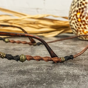 May include: A close-up of a brown and black glasses chain with brown and gold beads. The chain is made of braided cord and has a metal clasp.