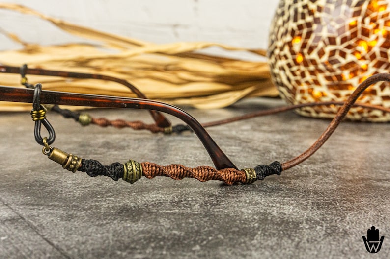 May include: A close-up of a brown and black glasses chain with brown and gold beads. The chain is made of braided cord and has a metal clasp.