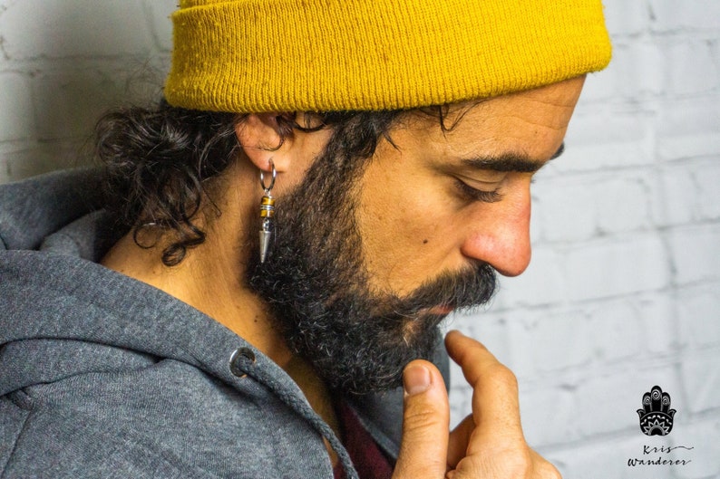 May include: A man with a beard wearing a yellow beanie and a gray hoodie with a silver hoop earring with a beaded dangle. The earring features a silver spike and brown beads.