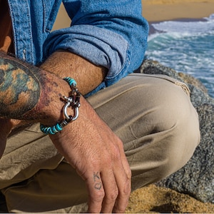Op de afbeelding: Een turquoise en bruine kralen armband met een zilveren sluiting. De armband wordt gedragen op de pols van een persoon.
